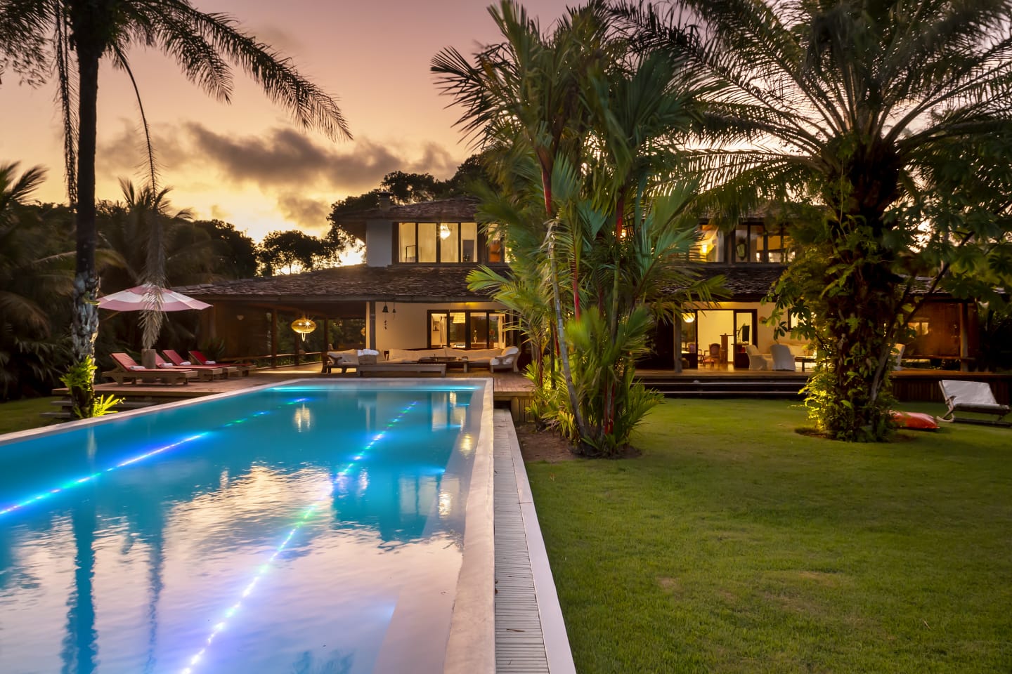 Casa cercada por jardim tropical, com piscina iluminada ao entardecer